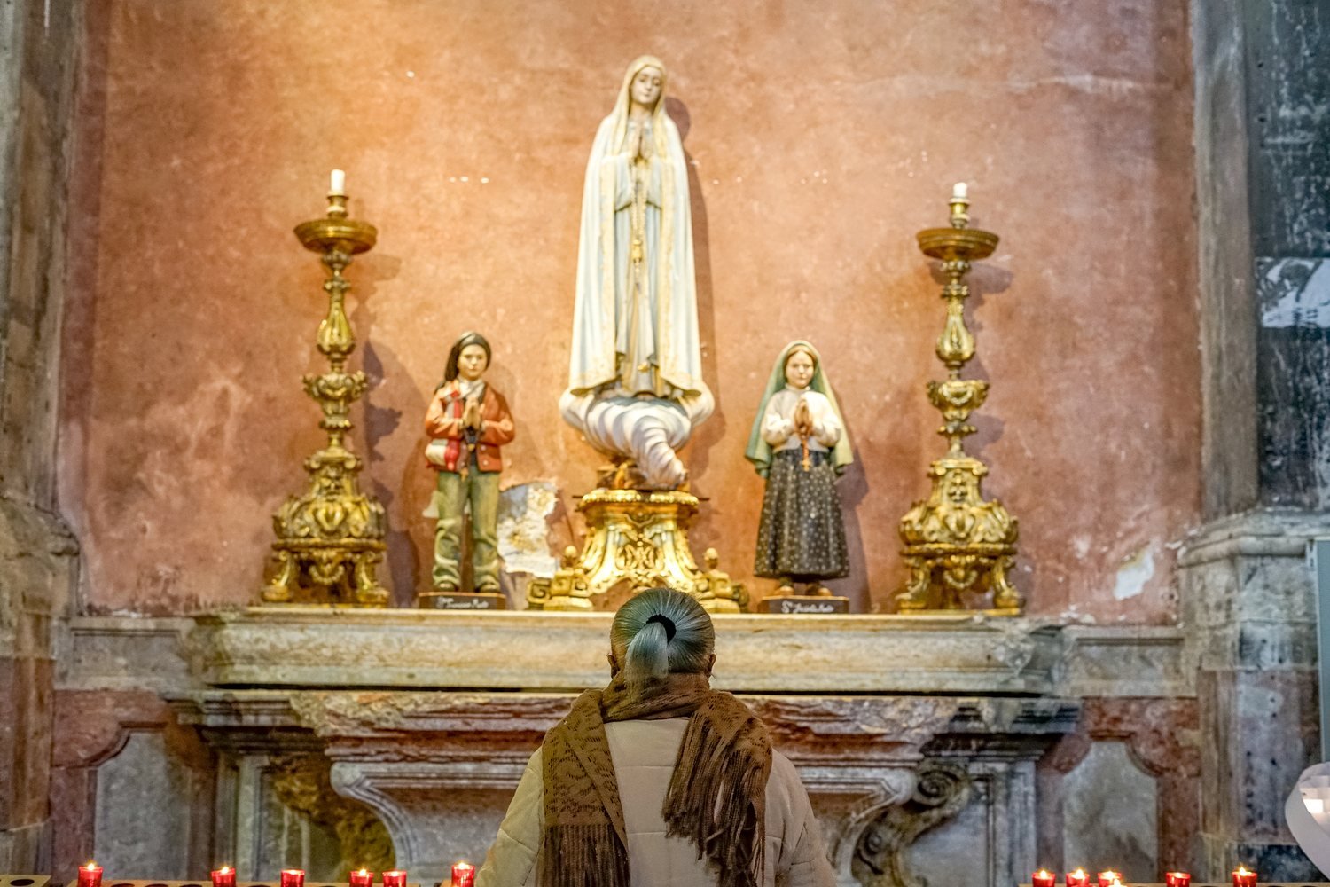 A woman kneeling in prayer before a statue of Our Lady, among lit candles